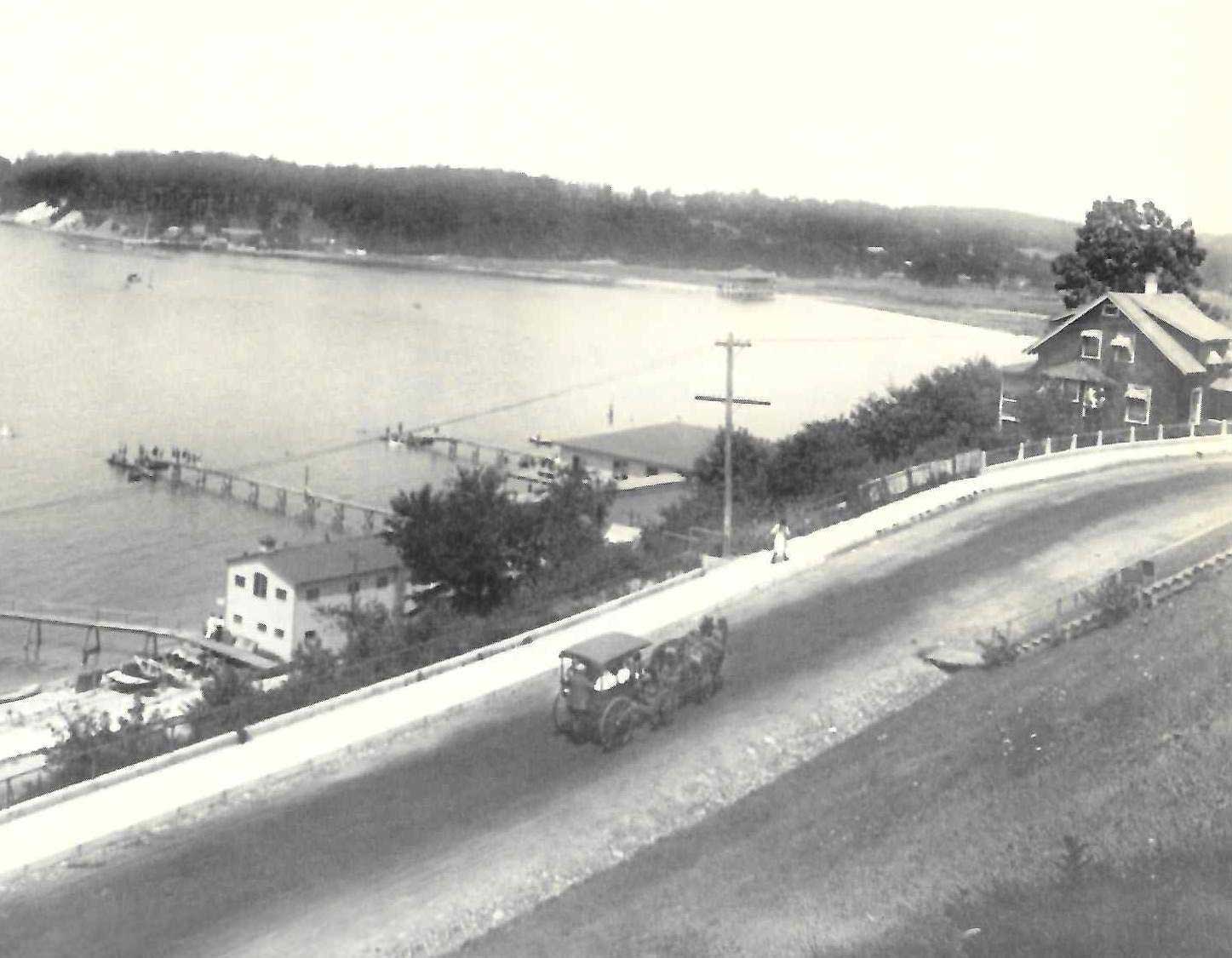 Hempstead-Harbor-1910 Black and white photo of a vintage car driving on a road beside a lake, with docks, a few buildings, and forested hills in the background.