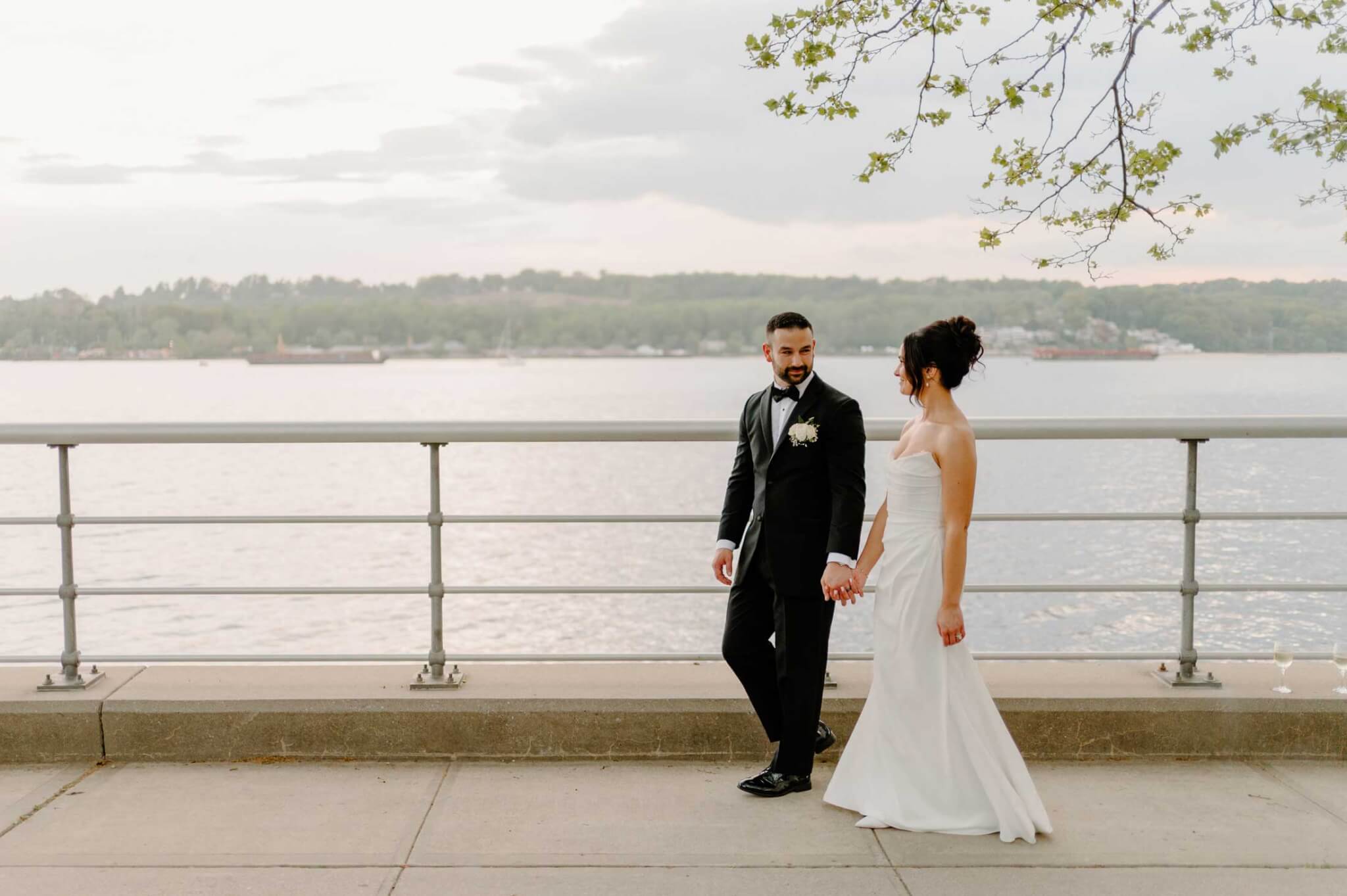 bride-and-groom-walk-along-sea-wall-by-sea-cliff-manor A man and woman in a suit holding hands.