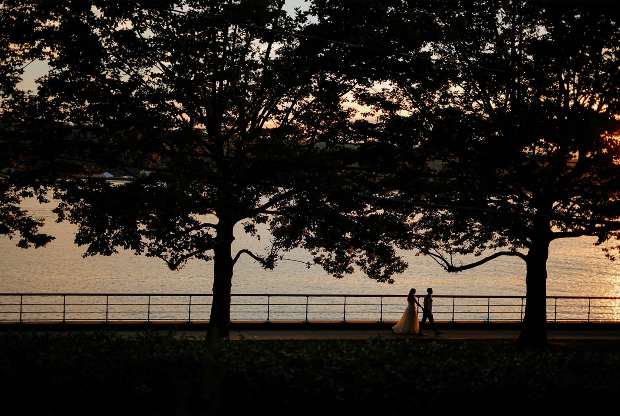 bride-and-groom-walking-along-sea-wall A silhouette of a couple walking on a sidewalk by a body of water.
