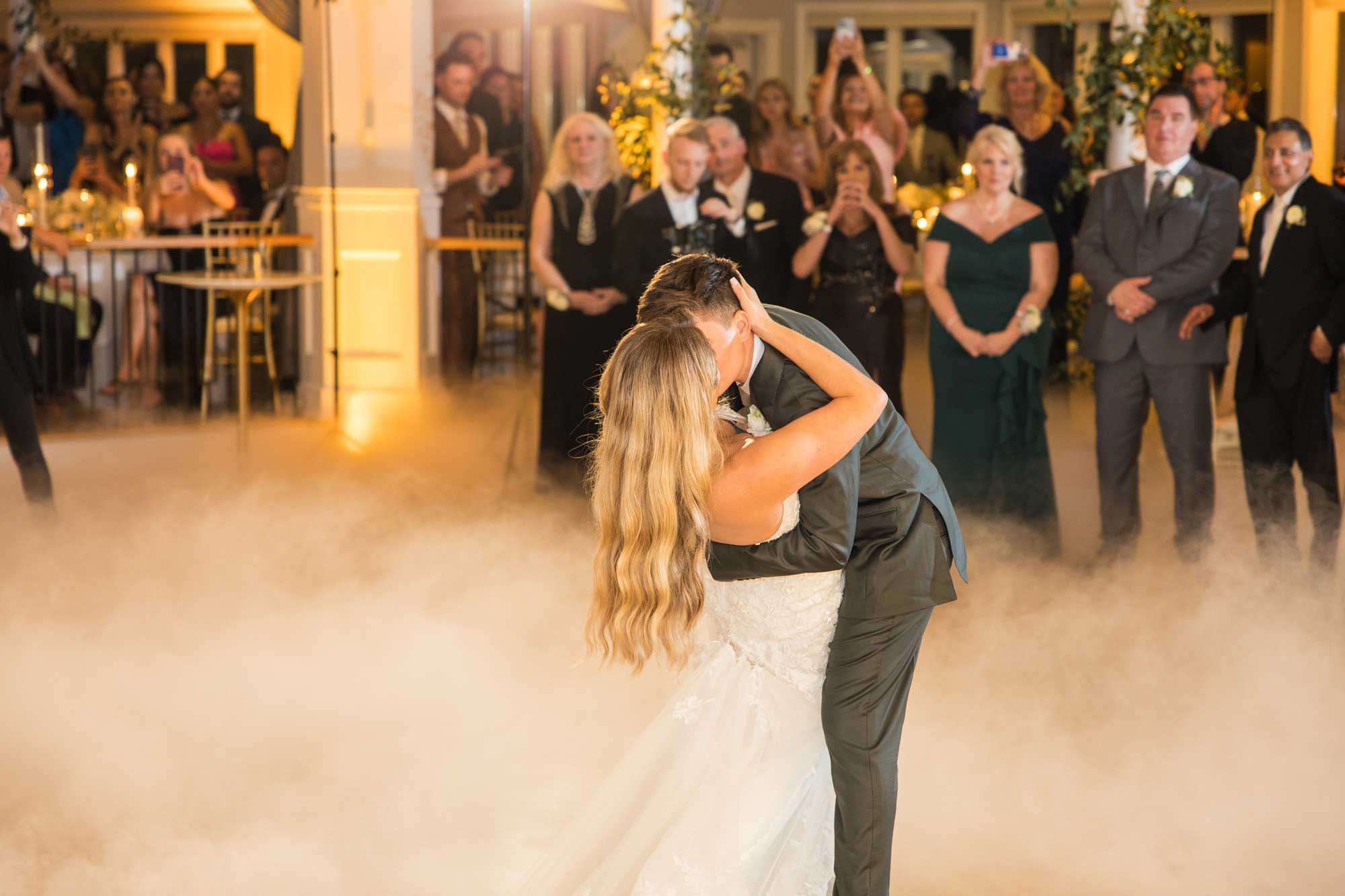 brides-wedding-dance-on-the-clouds-at-sea-cliff-manor A man and woman kissing in a room with people in the background.