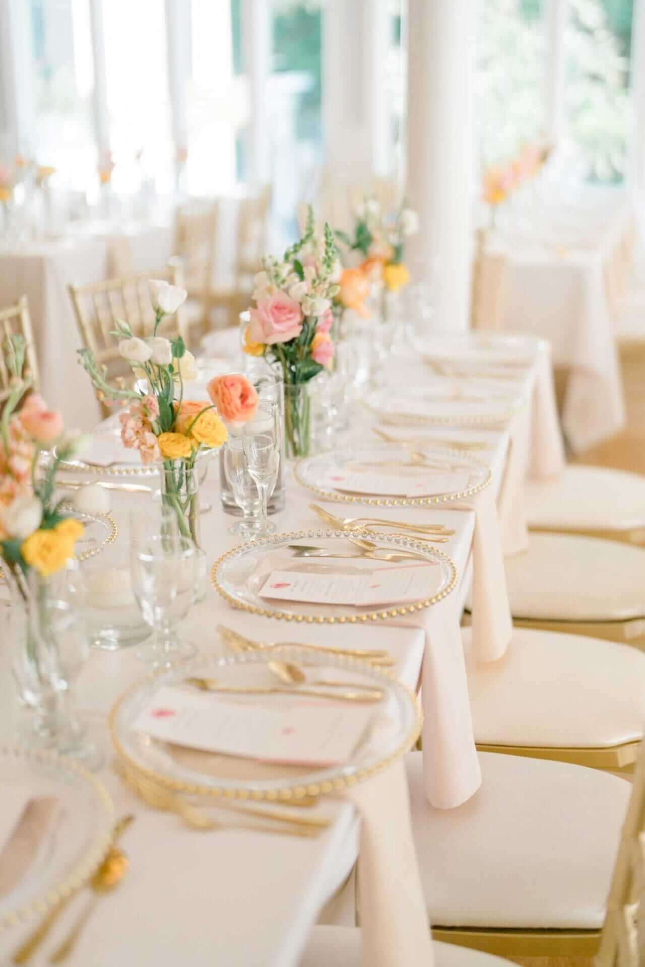 table-settings-with-menu-sea-cliff-manor Long banquet table set with gold-rimmed plates, gold flatware, glassware, and pastel floral centerpieces in a bright, airy room.