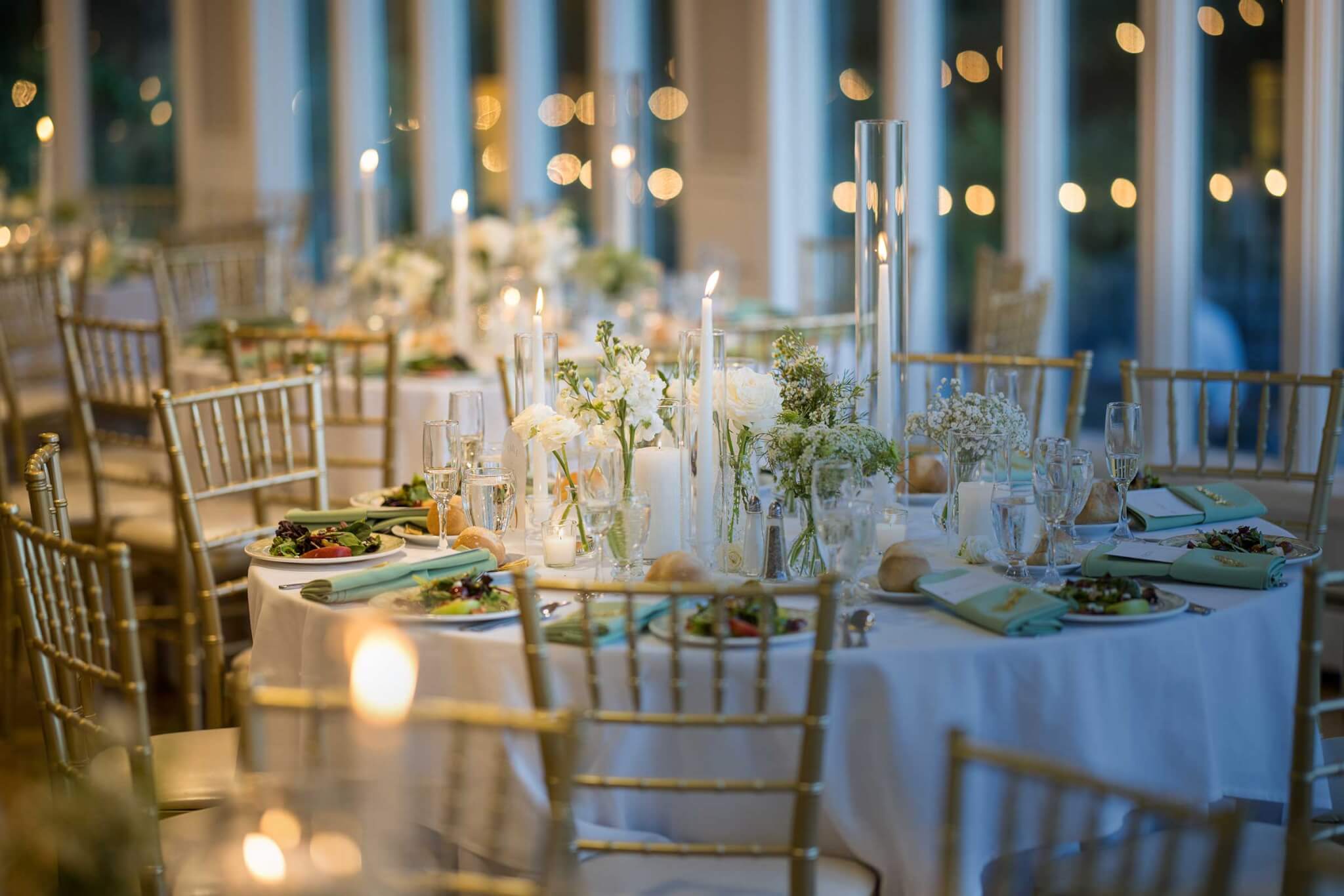 Elegant indoor banquet setting with round tables, gold chairs, white tablecloths, floral centerpieces, candles, and place settings prepared for a formal event.