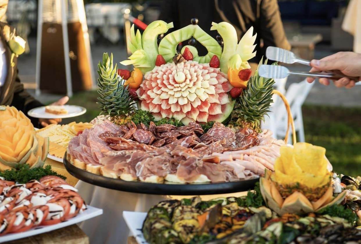 Chilled-Display Assorted cured meats, fruit carvings, and seafood displayed on a platter at an outdoor event, with people serving food.
