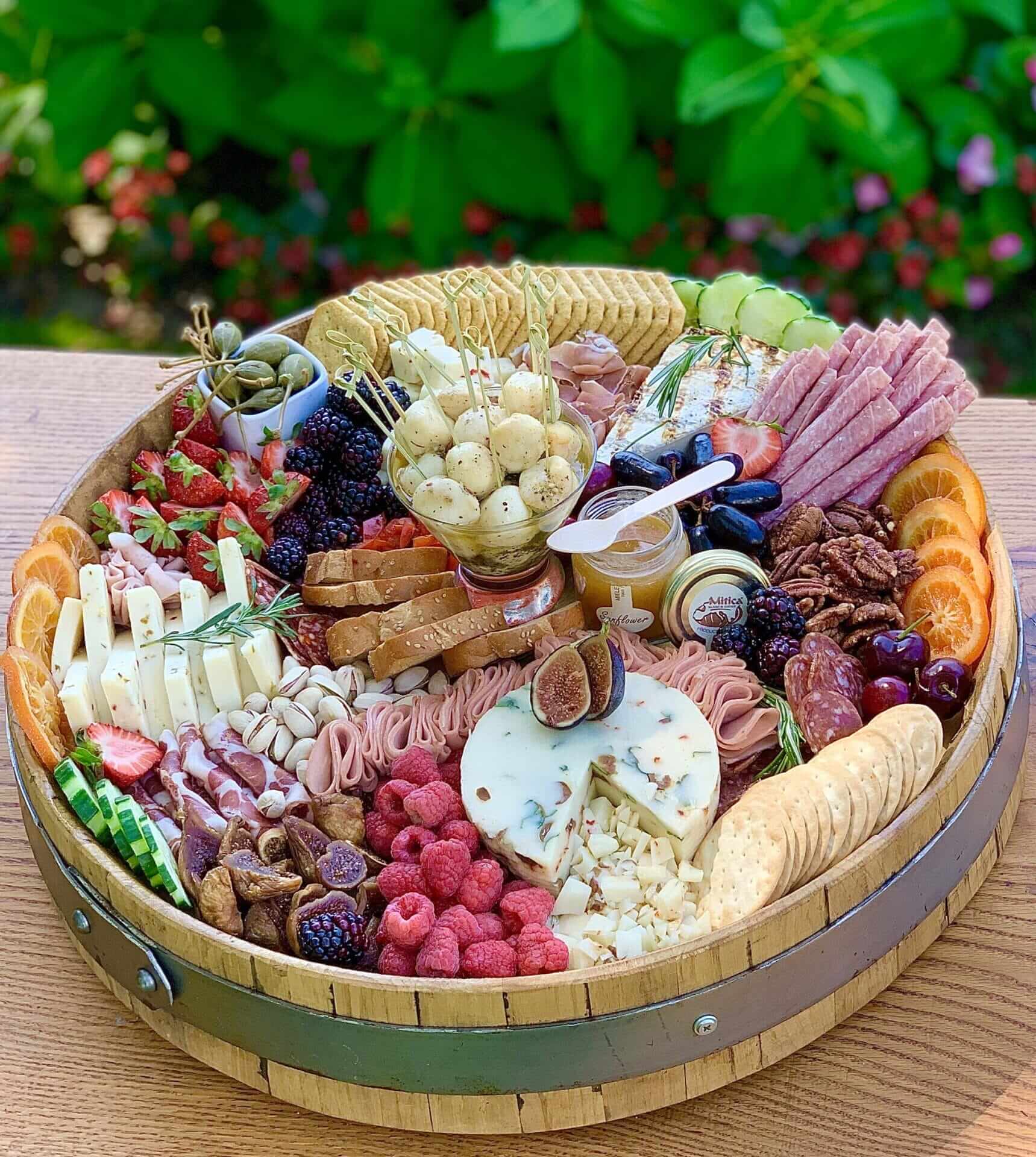 Sea-Cliff-Charcuterie-Board A large round charcuterie board with assorted cheeses, meats, crackers, nuts, fruits, olives, and spreads arranged neatly on a wooden surface outdoors.