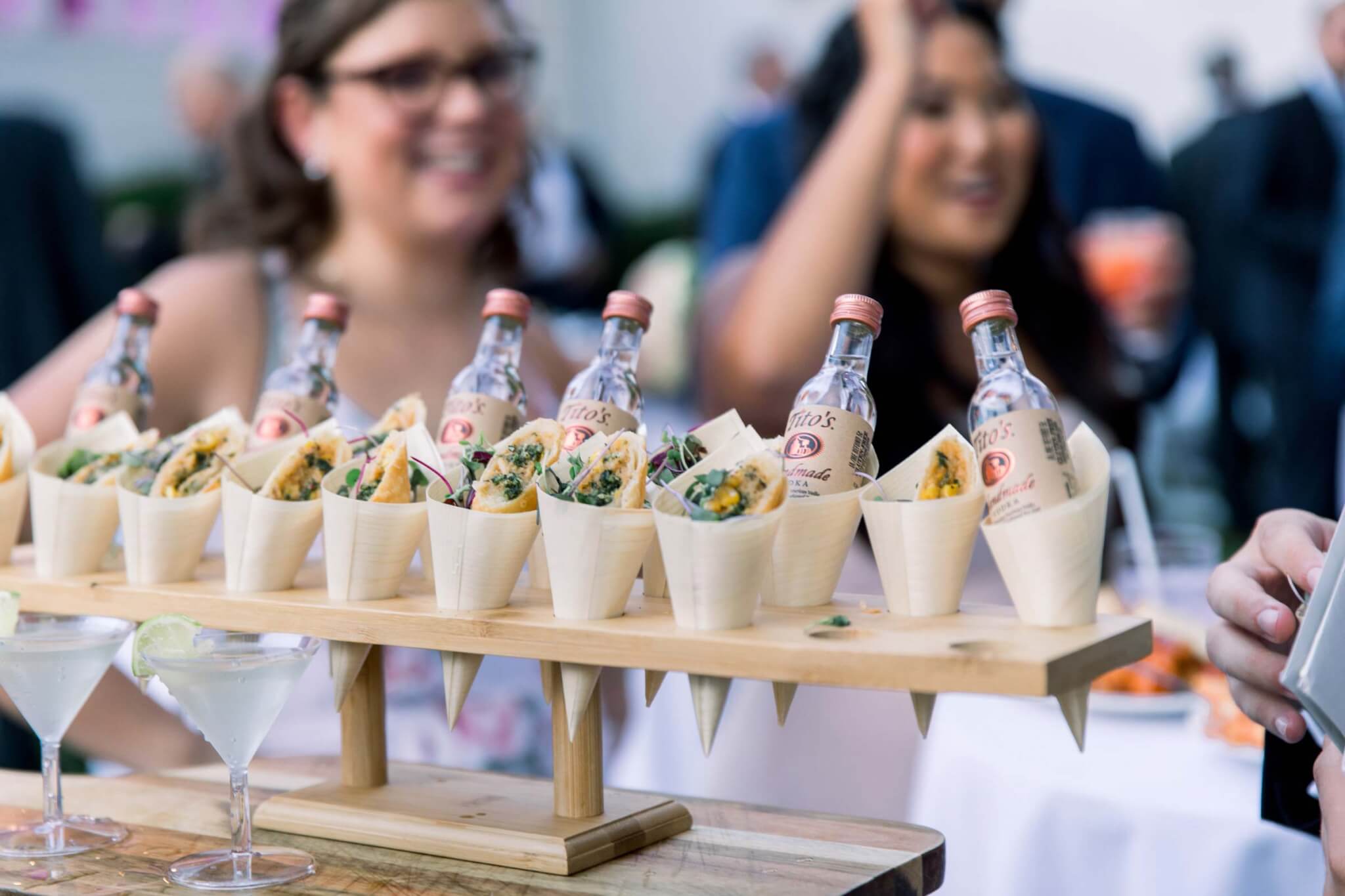 A wooden tray holds mini tacos and small bottles of Tito’s vodka in cones, with two women blurred in the background at an outdoor event.