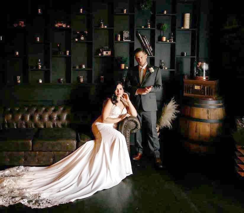bride-groom-rum-point-bar-sea-cliff-manor-brighter A bride in a white gown sits on a dark couch while a groom in a suit stands beside her in a dimly lit room with dark shelves and barrels.