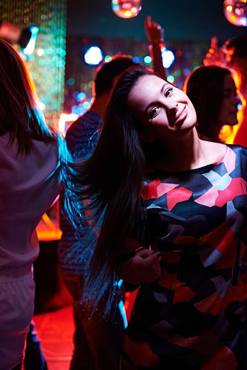 dancing-girl-186604757 Woman with long dark hair in a patterned dress smiles and poses at a nightclub with colorful lights and people dancing in the background.