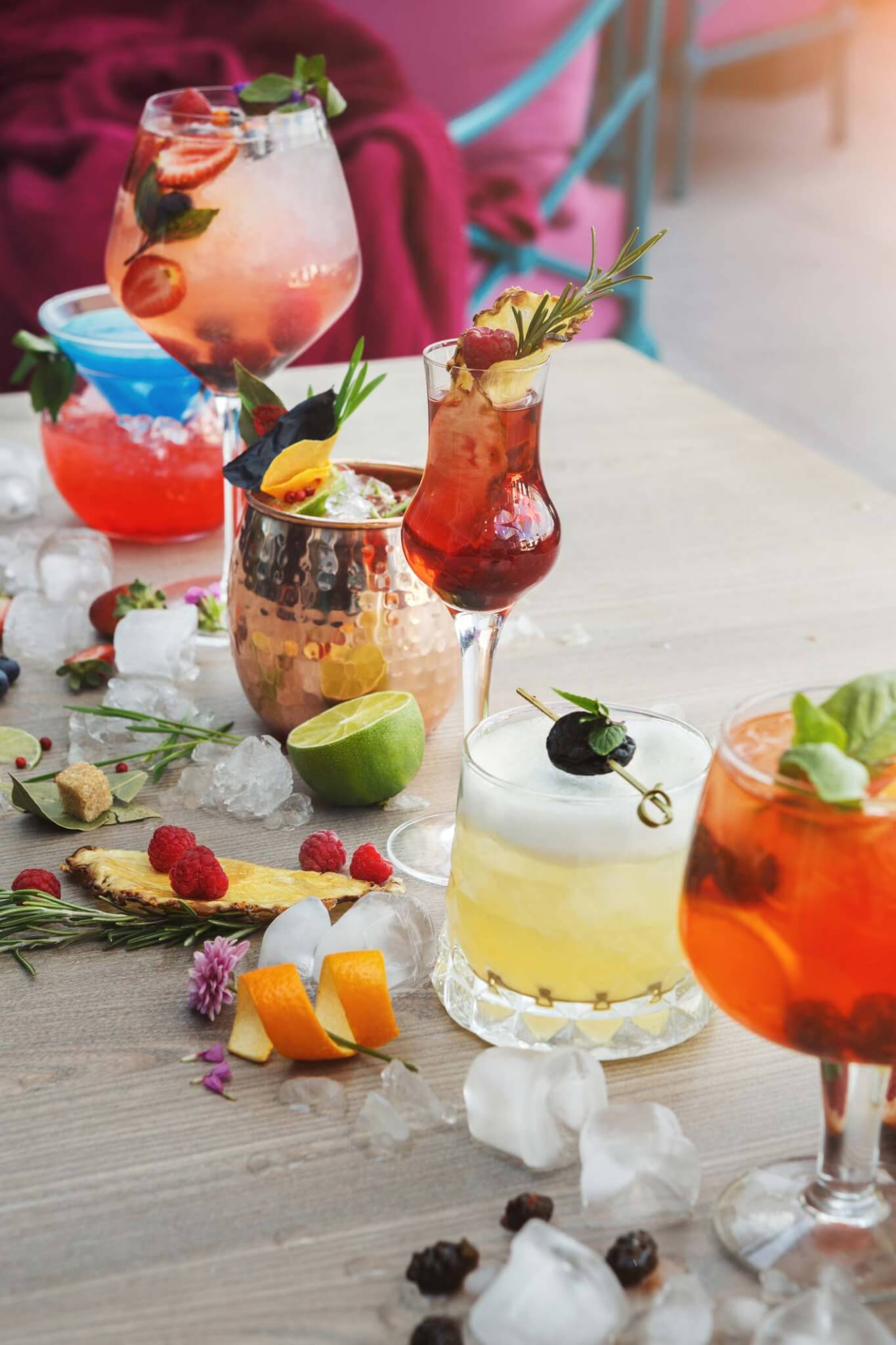 mocktails-cocktails-sea-cliff-manor-997698376 A variety of colorful cocktails garnished with fruit, herbs, and ice are arranged on a wooden table with scattered ice cubes, citrus slices, and berries.