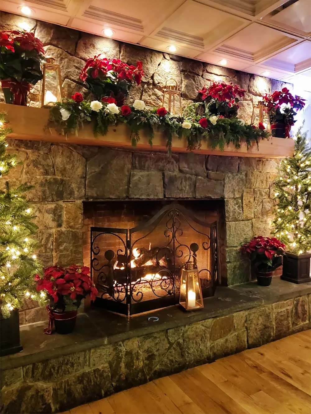 A stone fireplace with a lit fire, decorated with poinsettias, garlands, flowers, string lights, and lanterns, creating a festive holiday atmosphere.