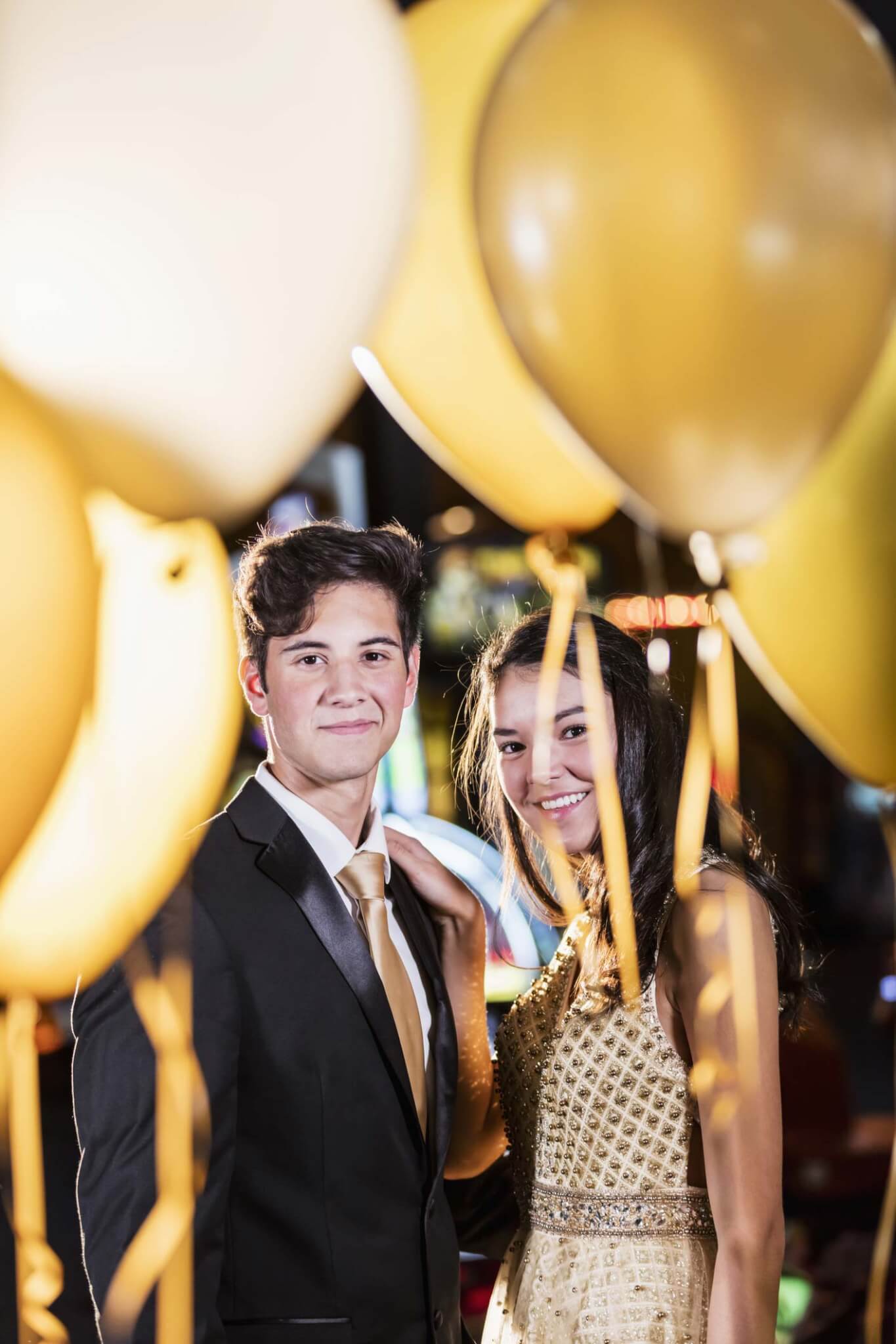 teen-boy-and-girl-balloons-1204280693 A young man in a suit and a young woman in a gold dress stand together, smiling, surrounded by gold and white balloons.