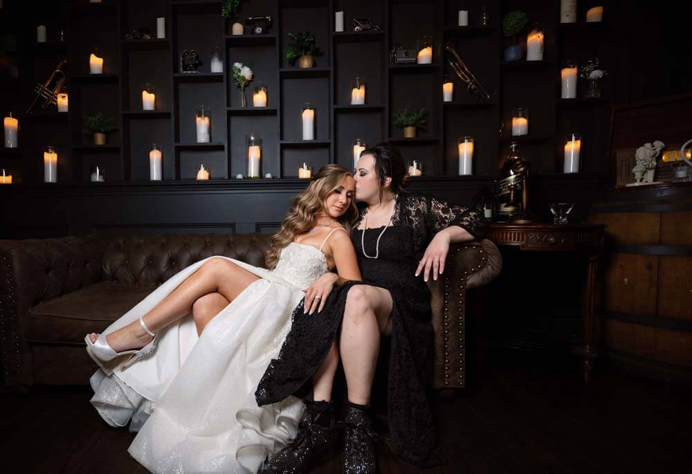 testimonial-jeanne-marie Two women in formal dresses sit closely on a couch; one in white with legs crossed, the other in black touches her partner's hair. Lit candles decorate shelves behind them.